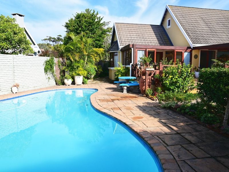 A well-maintained outdoor pool area featuring a curved swimming pool, paved patio surround, and a timber pergola attached to the main house structure.