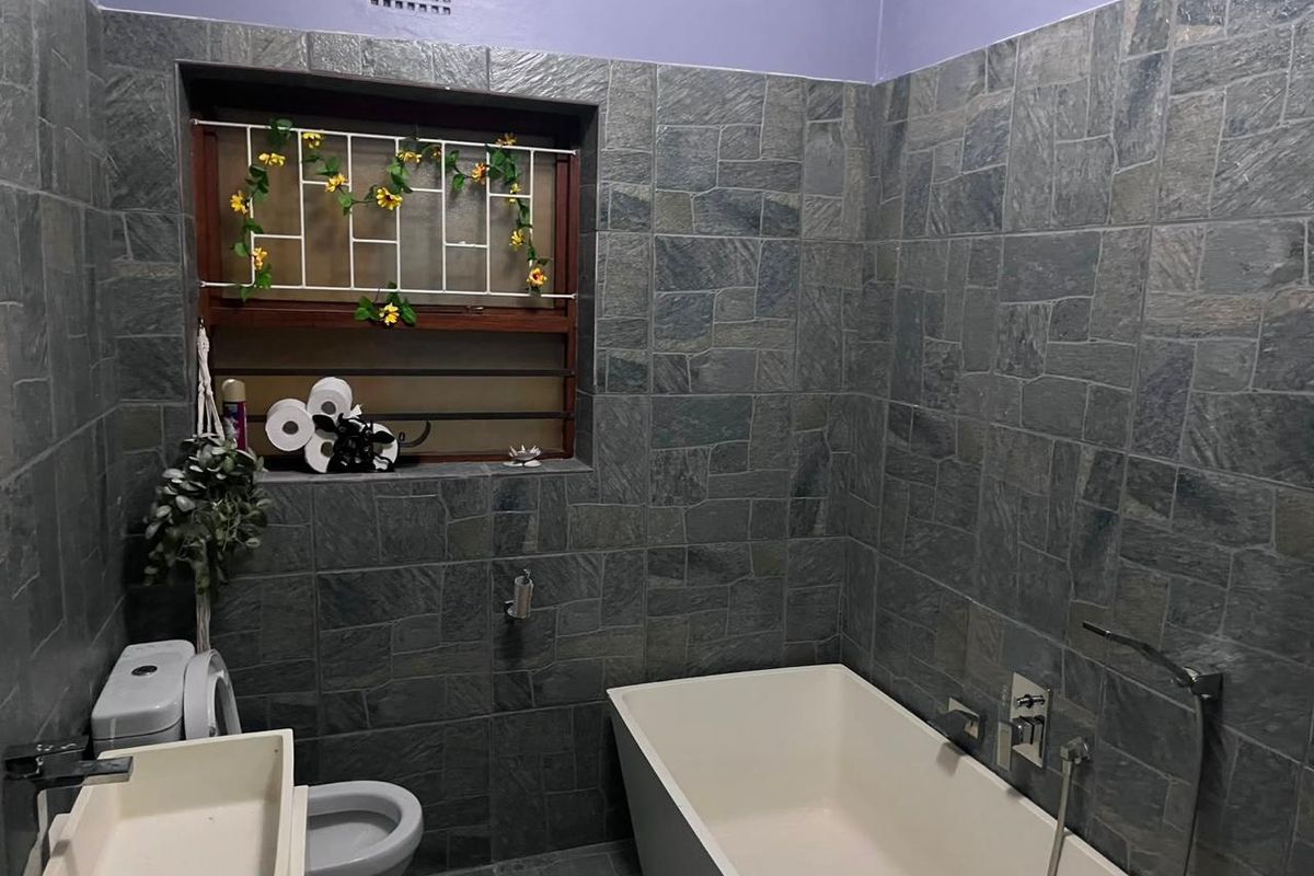 A bathroom featuring dark stone-effect wall tiling, a modern freestanding bathtub with handheld shower attachment, and a built-in vanity basin.