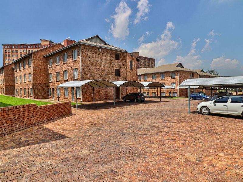 Exterior view of a multi-story brick apartment complex featuring a paved parking area with covered carports and well-maintained lawn spaces.