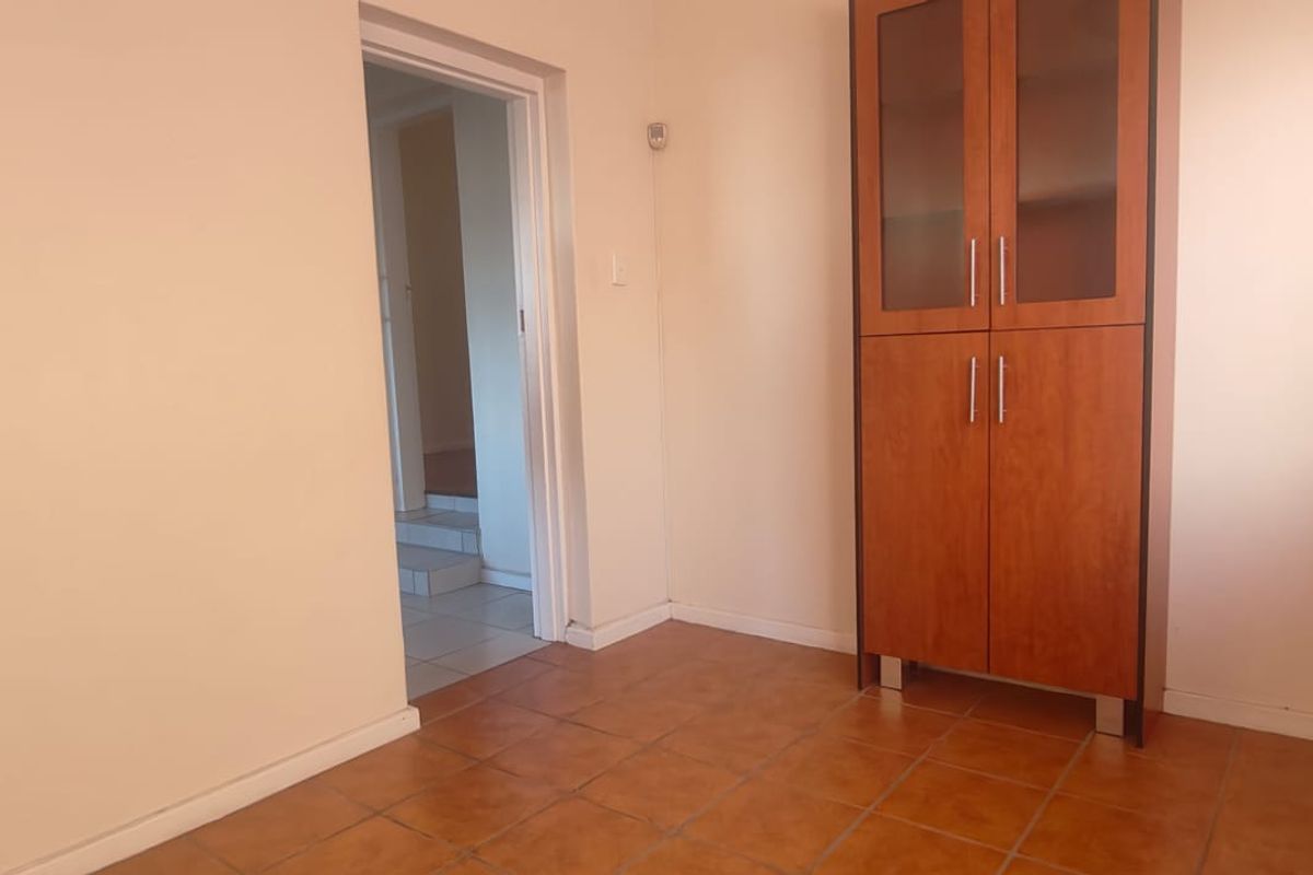 A simple, neutral living area featuring terracotta-style floor tiles and a built-in wooden storage cabinet, leading into an adjacent hallway.