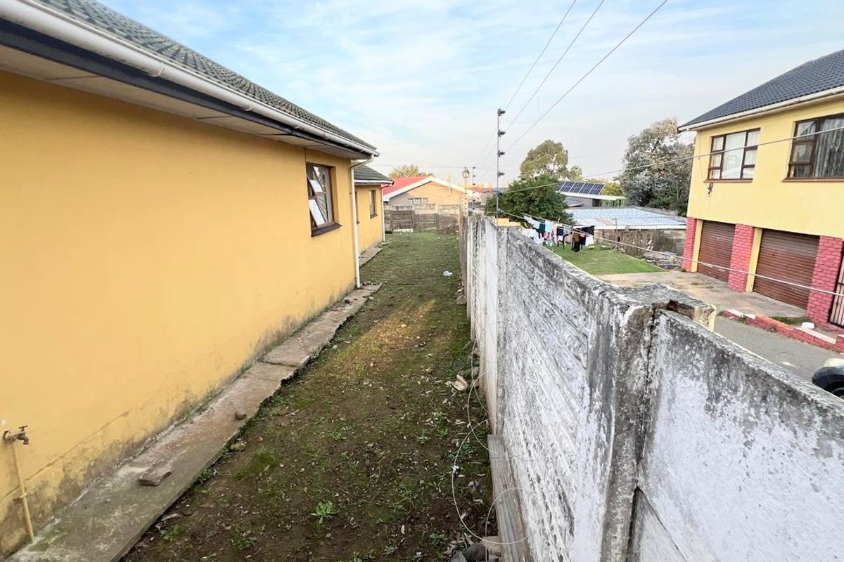 A narrow side yard area featuring a precast concrete boundary wall, concrete pathway, and exterior wall access with a water tap.