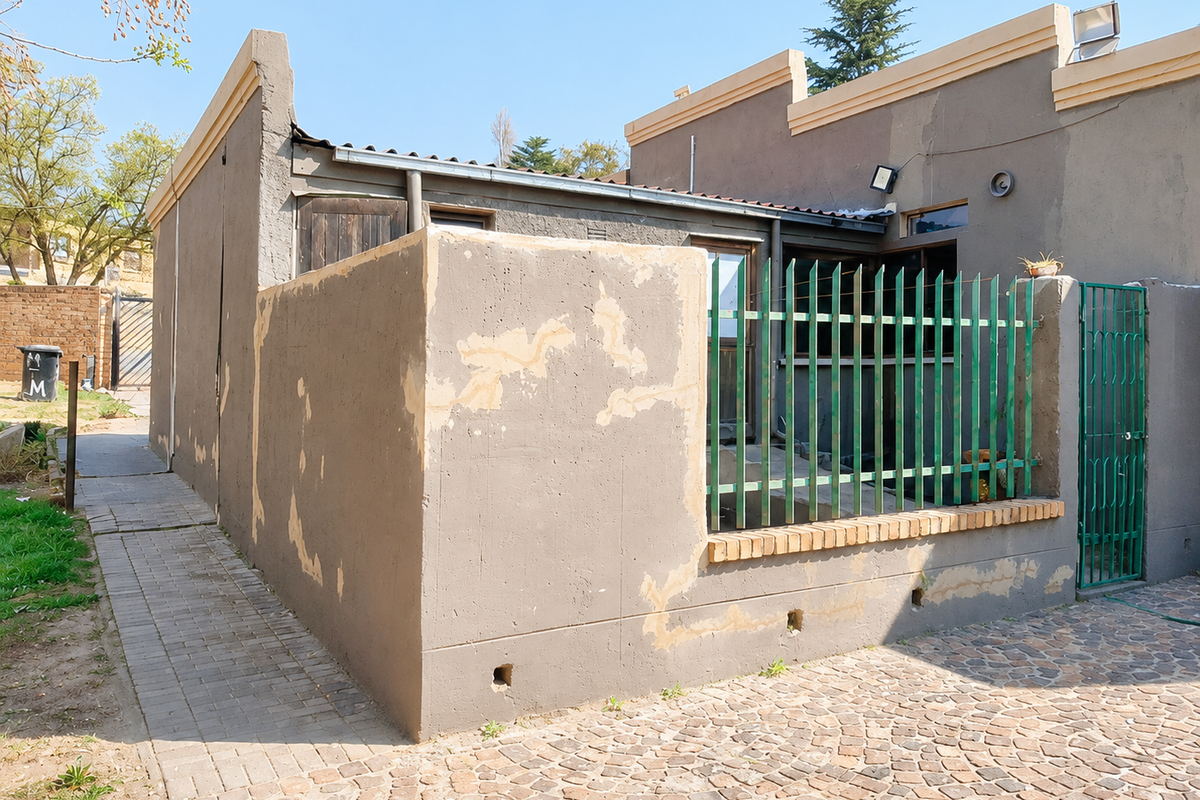 Rear exterior view of the property featuring a paved courtyard, metal security fencing, and a brick boundary wall with visible signs of exterior maintenance required.