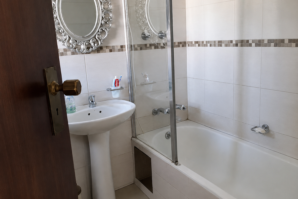 A functional bathroom featuring a pedestal basin, a bathtub with a glass shower screen, and neutral wall tiling with a decorative mosaic border.