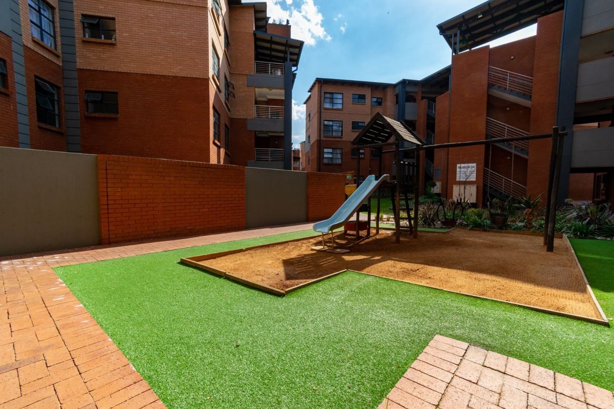 Communal outdoor area featuring a playground with a sand pit, artificial turf, and paved pathways situated between apartment blocks.