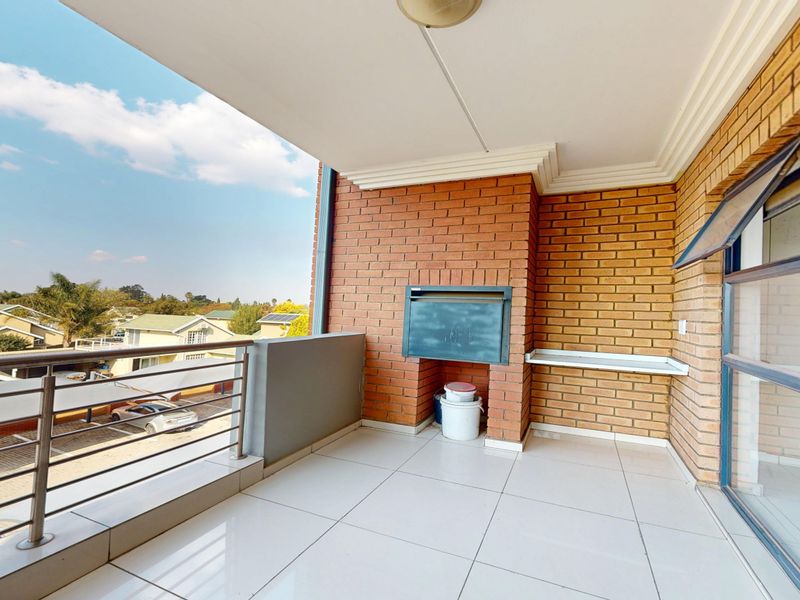 A spacious covered balcony featuring tiled flooring, a built-in braai, and a stainless steel balustrade with views of the surrounding complex.