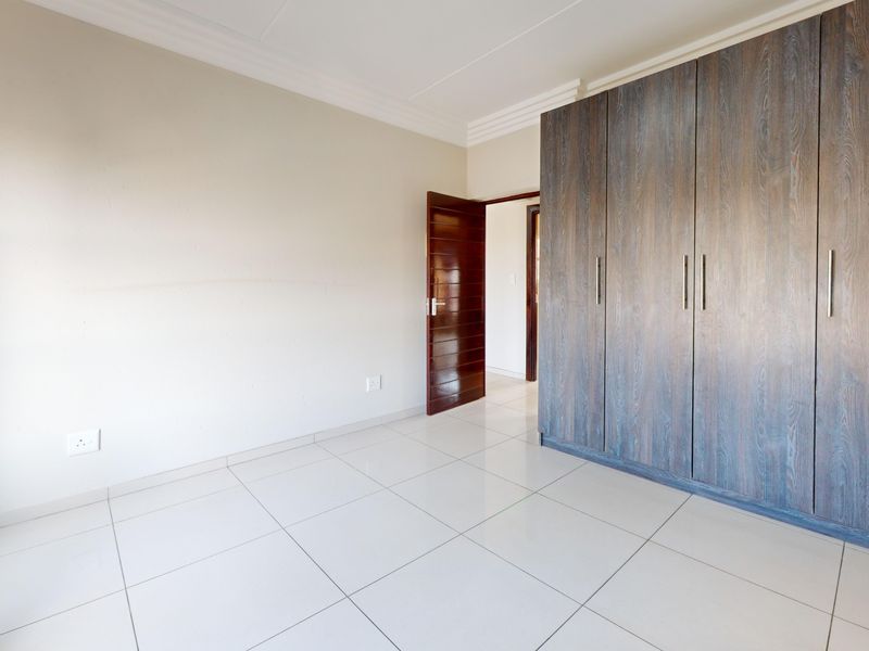 A bright, empty bedroom featuring neutral tiled flooring, white walls, and modern dark wood-finish built-in cupboards.