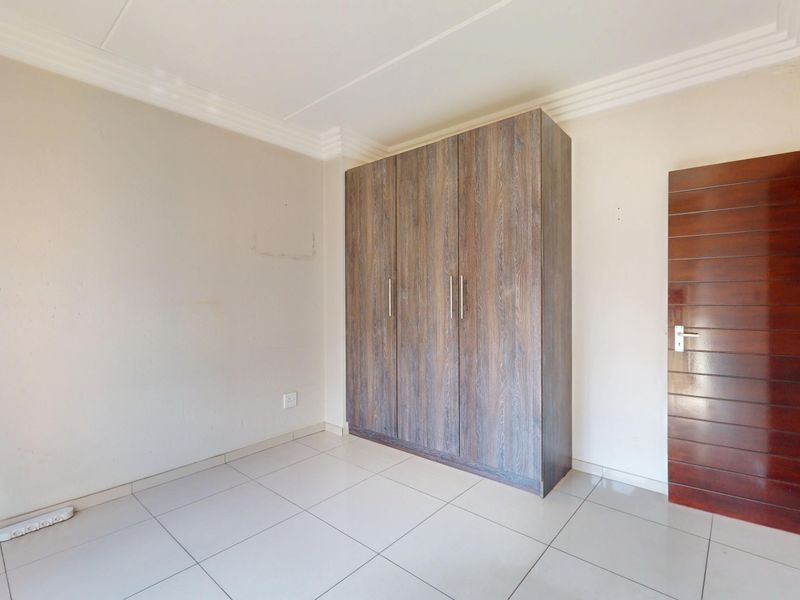 A clean, empty bedroom featuring neutral tiled flooring, a built-in wooden wardrobe, and a solid timber door.
