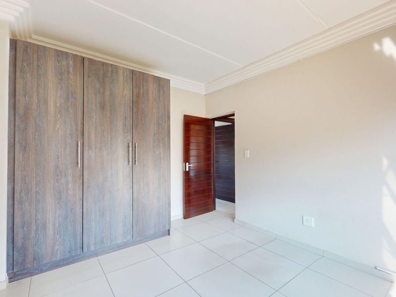 A bright, empty bedroom featuring neutral tiled flooring, built-in wooden cupboards, and a solid wood door, offering a clean and versatile living space.