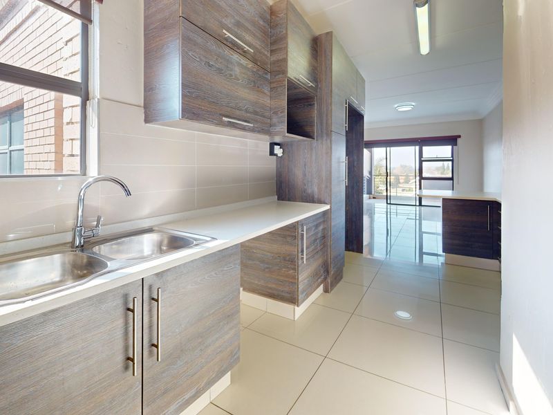 A clean, modern kitchen featuring wood-look laminate cabinetry, stainless steel sink, tiled splashback, and neutral floor tiles, leading into an open-plan living area.