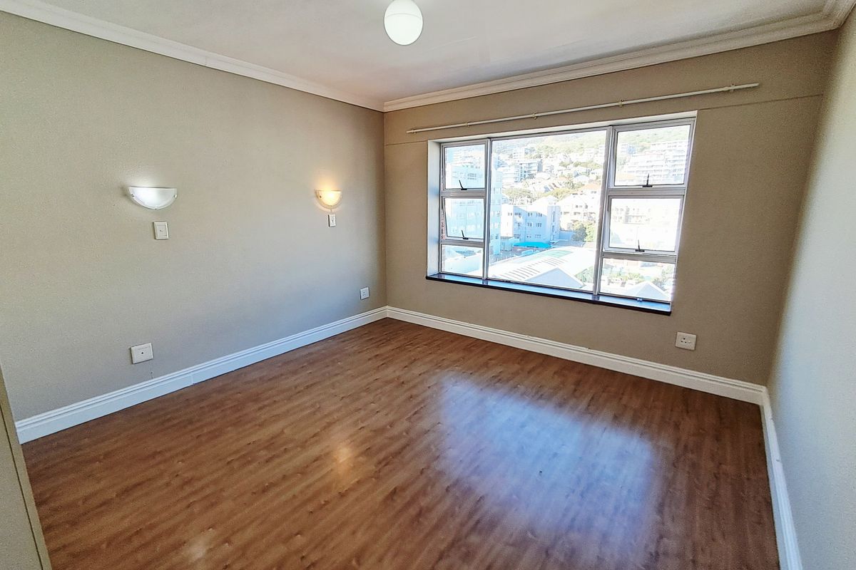A bright, empty bedroom featuring wood-look laminate flooring, wall-mounted sconce lighting, and large windows offering an urban view.