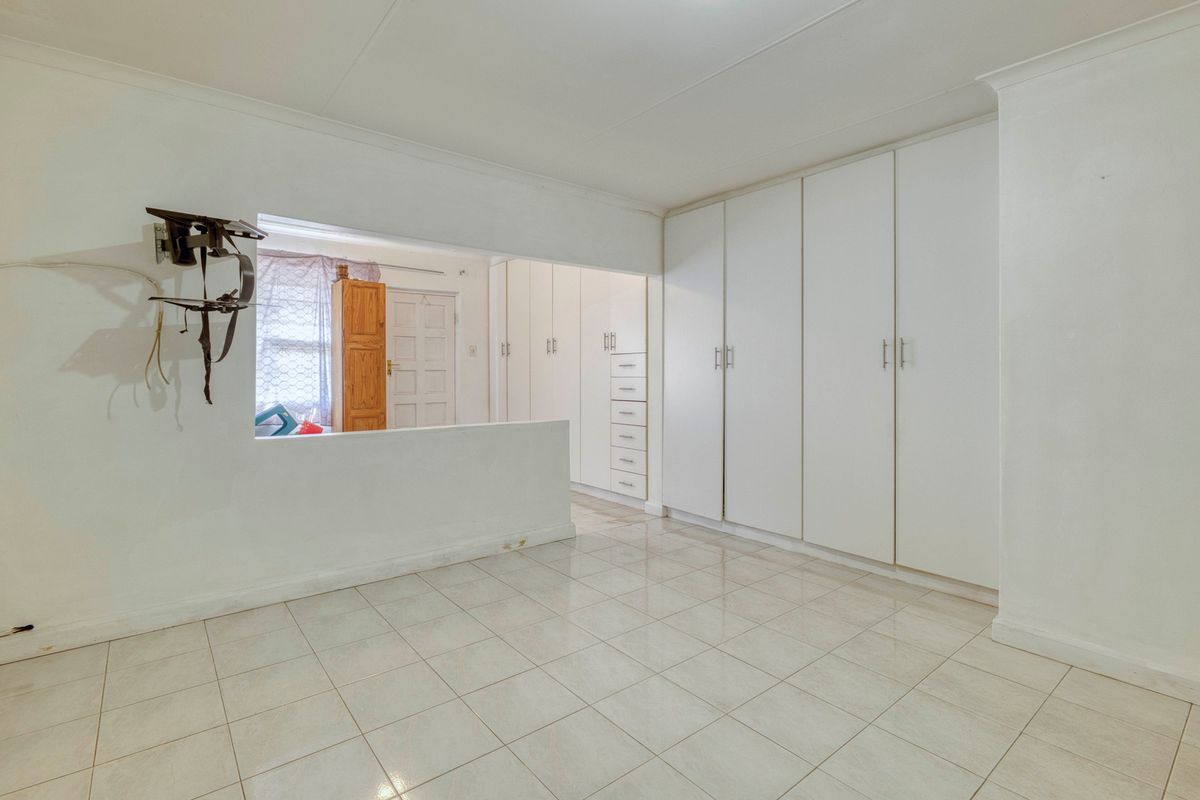 A spacious, empty bedroom featuring extensive built-in cupboards, neutral tiled flooring, and a semi-open partition wall leading to an adjacent area.