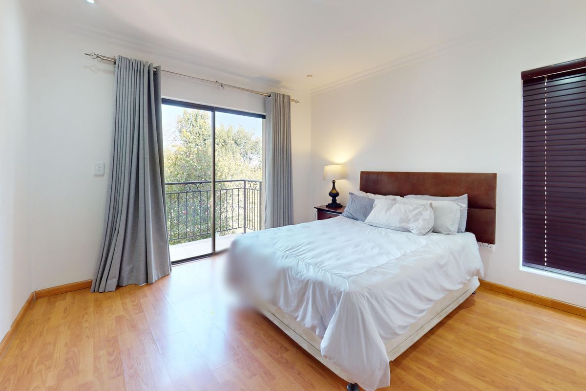 A well-lit bedroom featuring laminate flooring, a sliding glass door leading to a balcony, and dark venetian blinds.