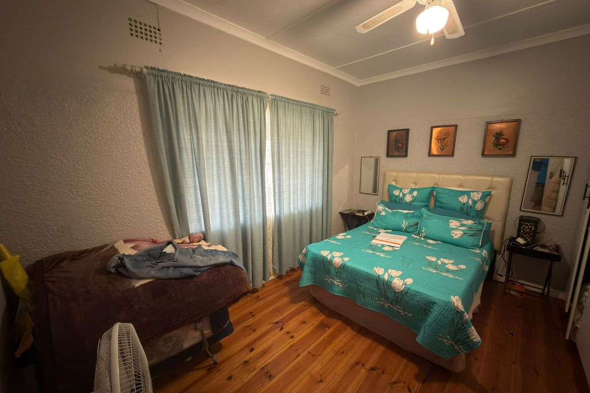 A bedroom featuring original timber flooring, a ceiling fan, and large windows with curtains. The room shows signs of ceiling wear.