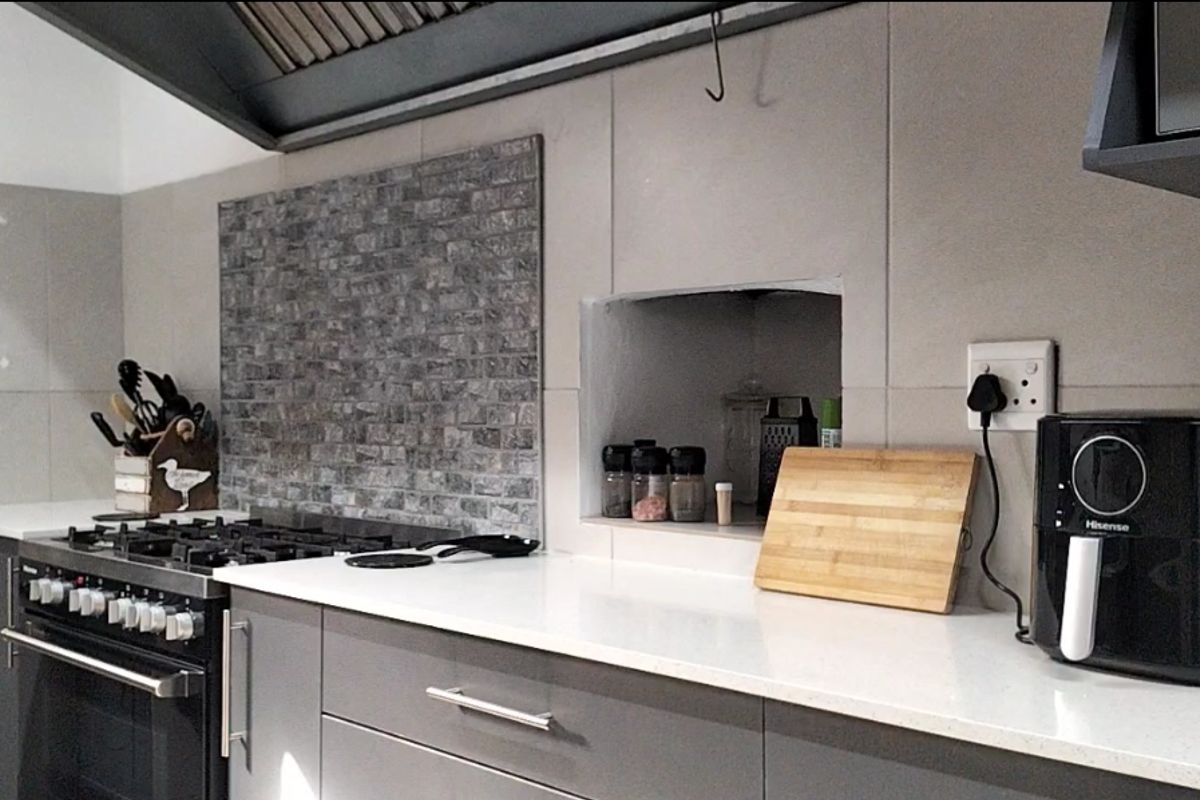 Modern kitchen featuring grey cabinetry, white stone countertops, a gas stove with extractor fan, and a decorative brick-style tiled backsplash.