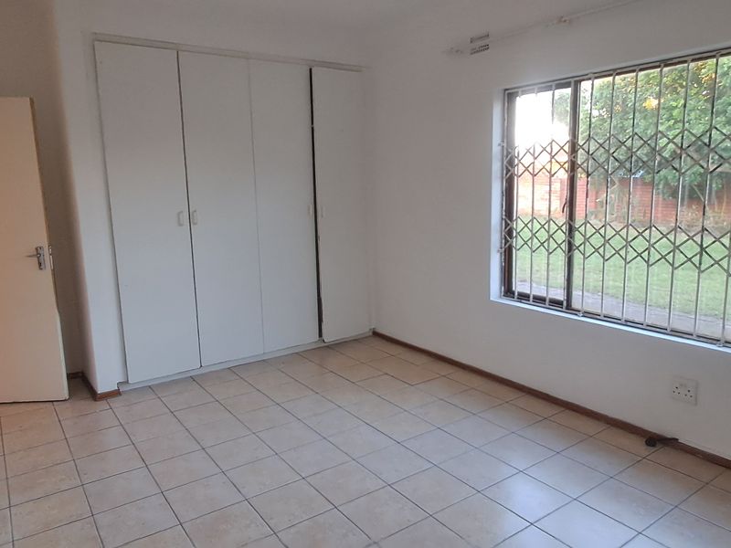 A bright, empty bedroom featuring tiled flooring, built-in cupboards, and a window fitted with a security gate.