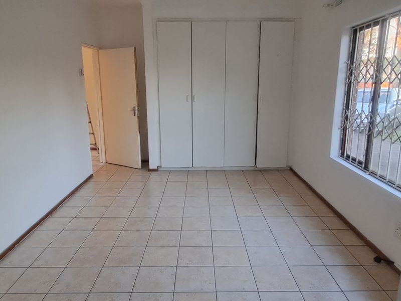 A bright, empty bedroom featuring neutral tiled flooring, built-in cupboards, and security bars on the window.