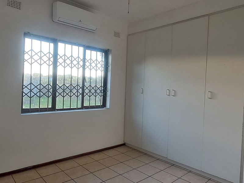 A clean, empty bedroom featuring built-in cupboards, tiled flooring, and a wall-mounted air conditioning unit with security bars on the window.