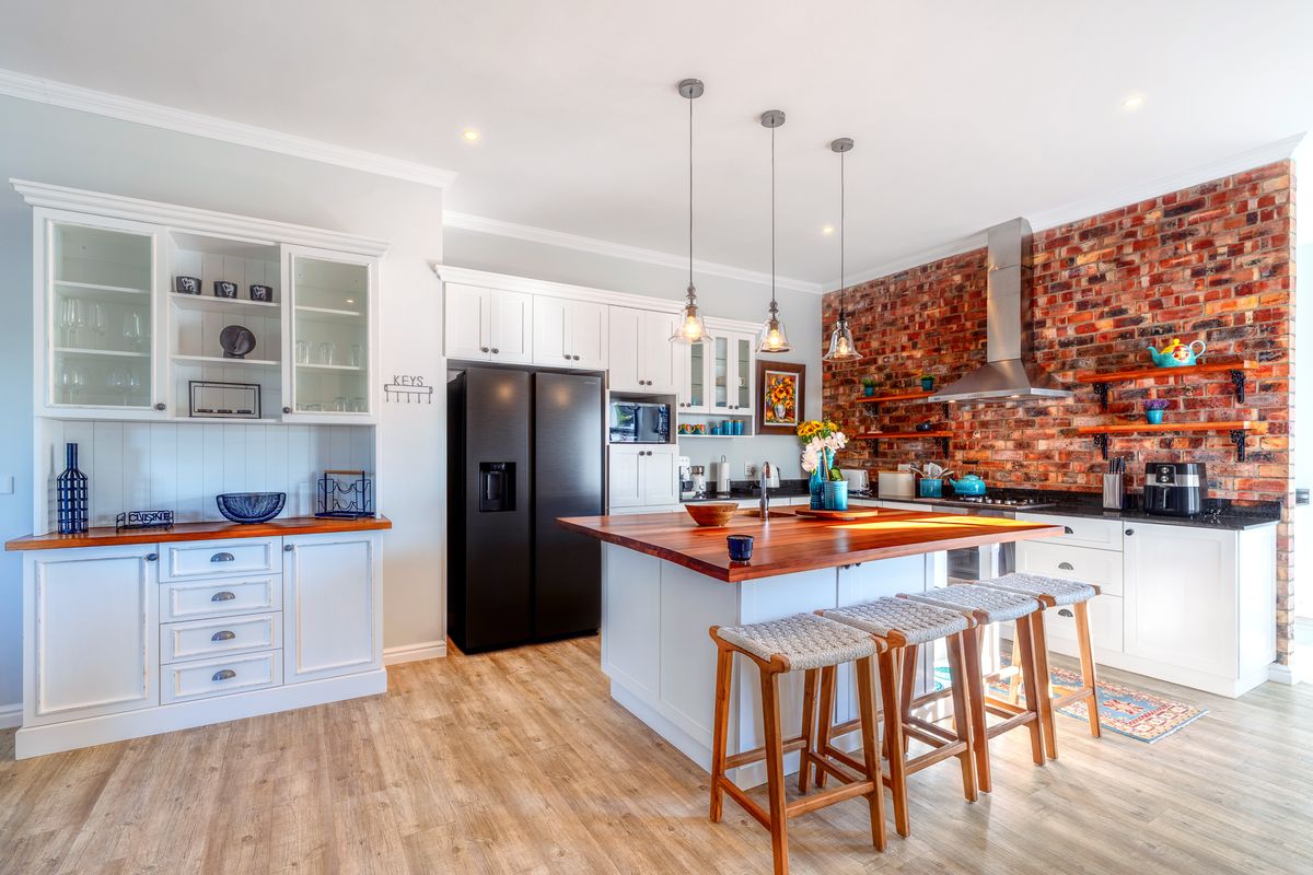 Modern kitchen featuring a large island with wooden countertop, exposed brick feature wall, white cabinetry, and high-quality pendant lighting.