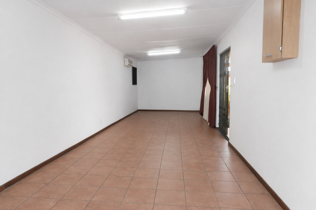 An empty, open-plan living area featuring tiled flooring, fluorescent lighting, a wall-mounted air conditioning unit, and a security gate leading to the exterior.