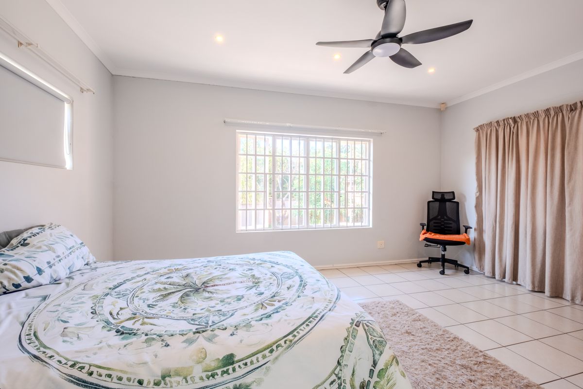 A spacious bedroom featuring neutral tiled flooring, a modern ceiling fan, recessed lighting, and a window fitted with security bars.