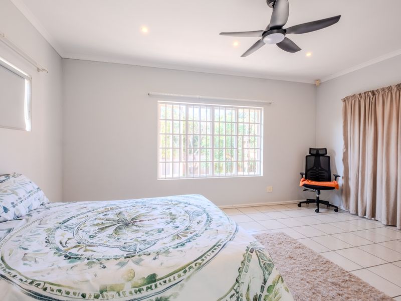 A spacious bedroom featuring neutral tiled flooring, a modern ceiling fan, recessed lighting, and a window fitted with security bars.