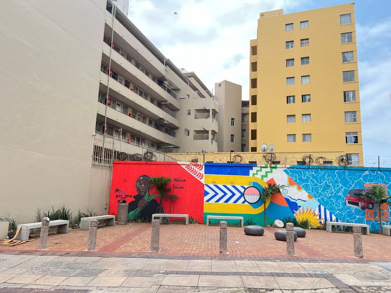 A paved outdoor courtyard area featuring decorative mural walls, concrete seating, and security fencing between apartment buildings.