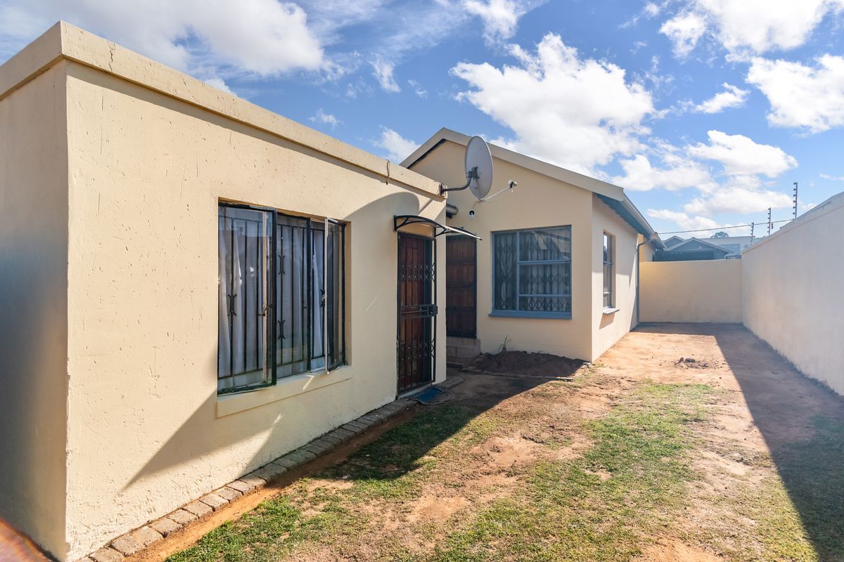 Rear exterior view of the property featuring a paved walkway, security gates, and a boundary wall with a small garden area.