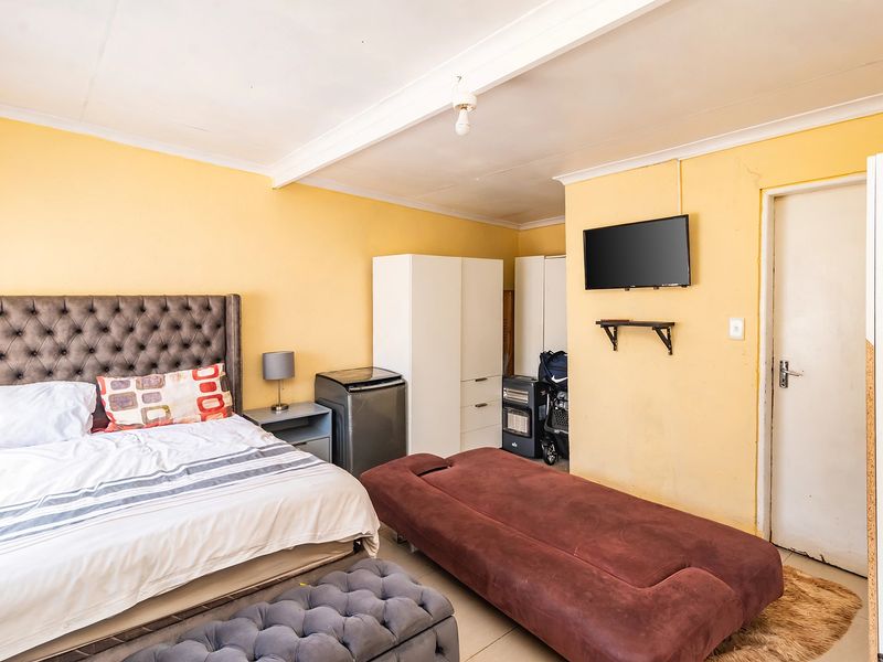 A bedroom featuring tiled flooring, built-in wardrobe storage, and a wall-mounted television, with yellow painted walls and an exposed ceiling beam.