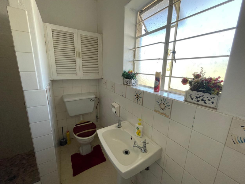 A compact bathroom featuring a wall-mounted basin, toilet, and louvered storage cabinet, finished with tiled walls and a window with security bars.