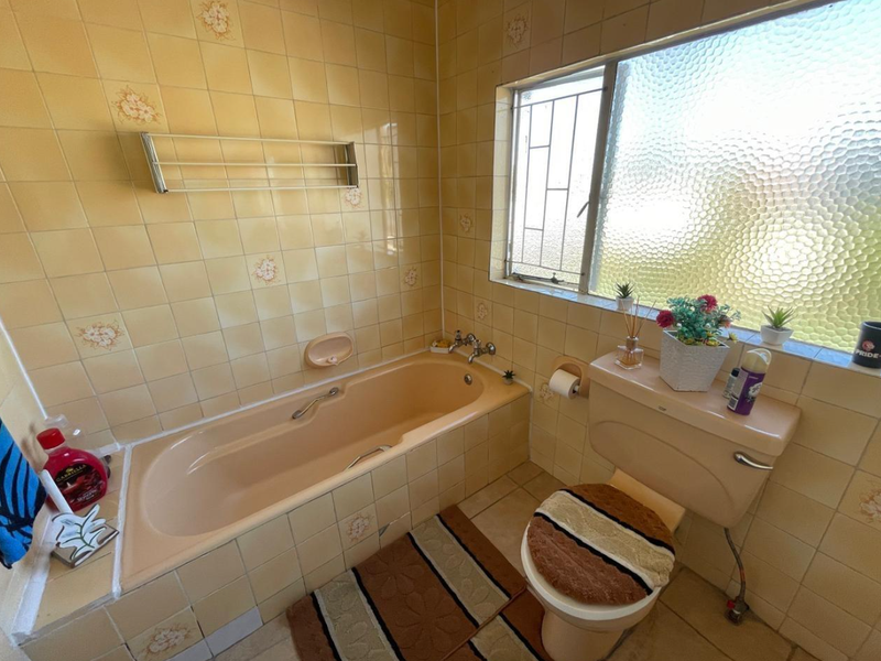 A dated bathroom featuring original peach-toned sanitaryware, yellow floral wall tiles, and a window with security bars.