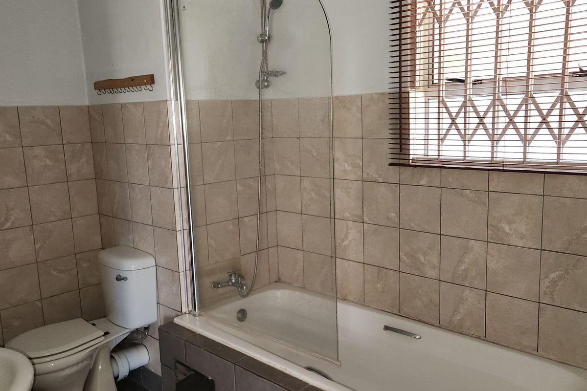 A functional bathroom featuring a tiled bathtub with a glass shower screen, a standard toilet, and a window with security bars.
