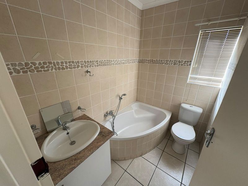 A functional bathroom featuring a corner bathtub, vanity basin, and toilet, finished with neutral wall and floor tiling.