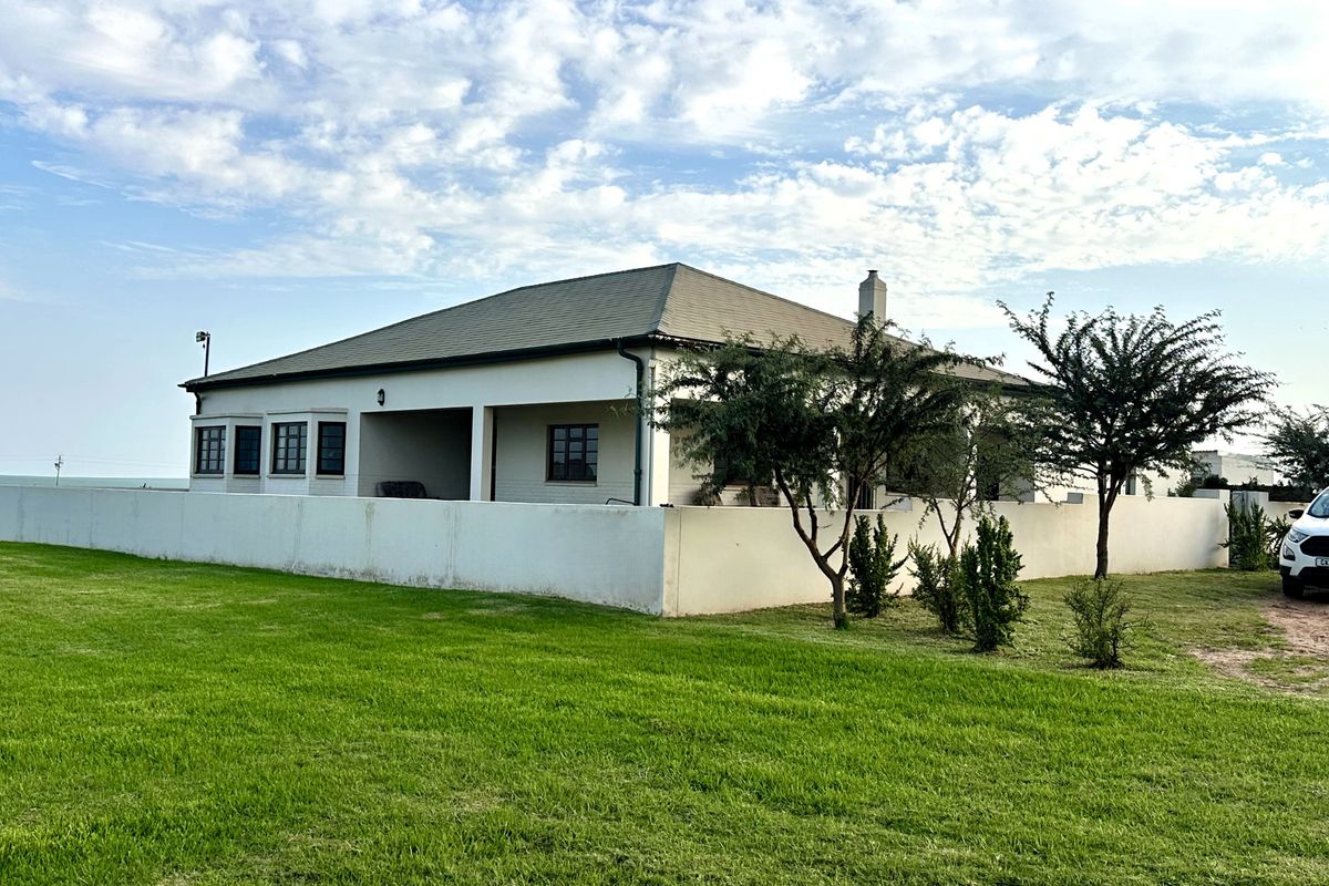 A well-maintained single-story house featuring a hipped roof, chimney, and a covered patio area, set within a spacious garden with a perimeter wall.