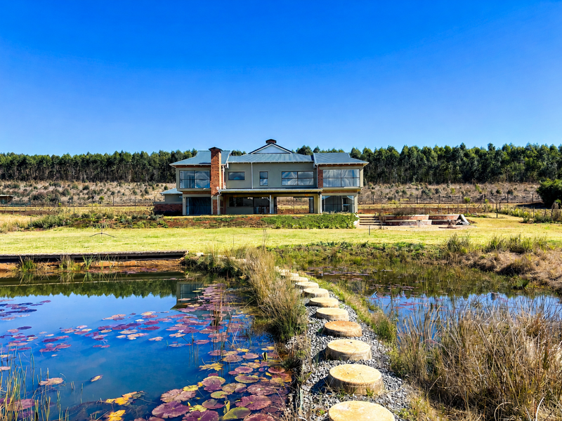 A rear view of a modern two-story house featuring a brick chimney, expansive windows, and a covered patio overlooking a natural pond and landscaped garden.