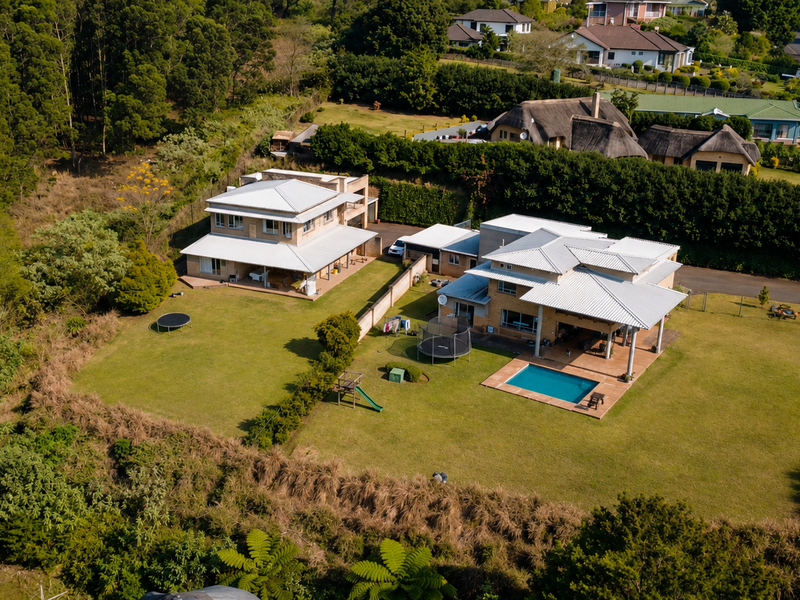 Aerial view of a residential property featuring two main dwellings, a swimming pool, expansive lawns, and established garden surroundings.