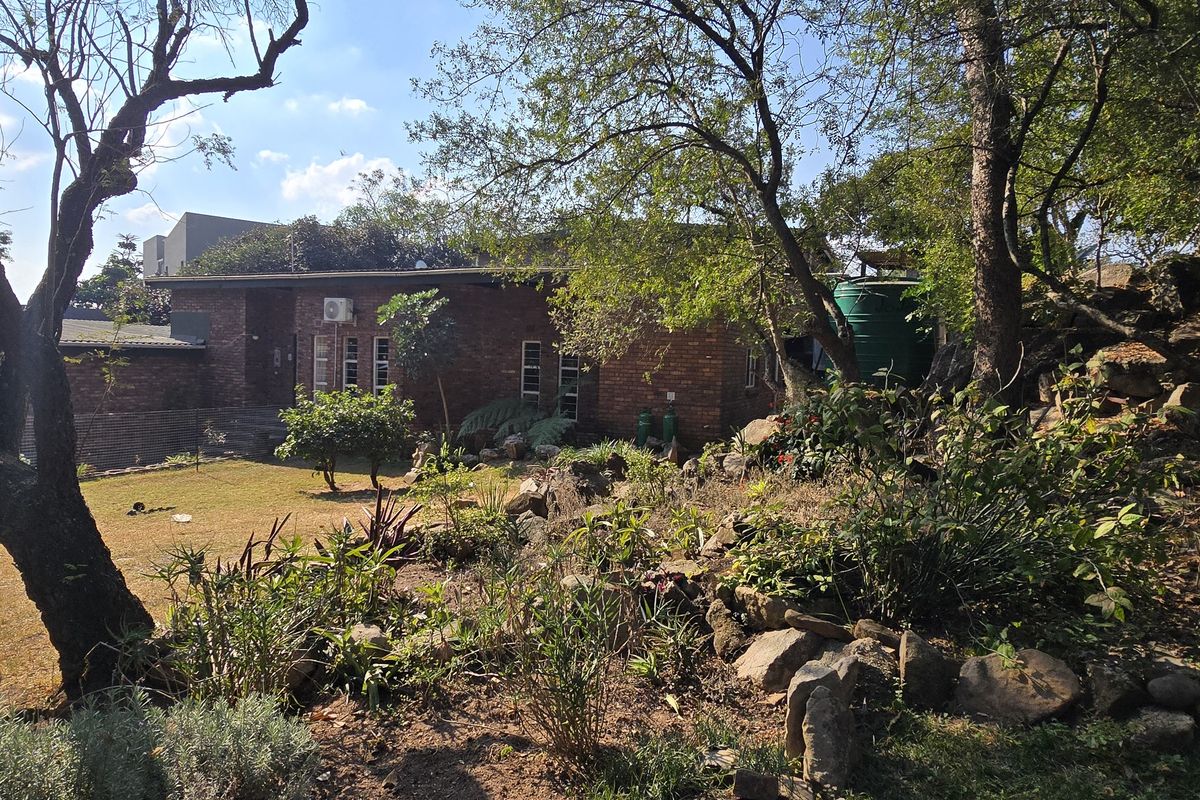 A view of the rear brick facade of the house, featuring a landscaped garden with rockery, mature trees, and a water storage tank.