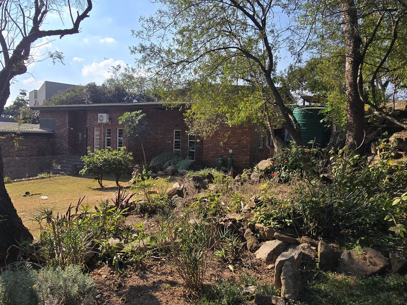 A view of the rear brick facade of the house, featuring a landscaped garden with rockery, mature trees, and a water storage tank.