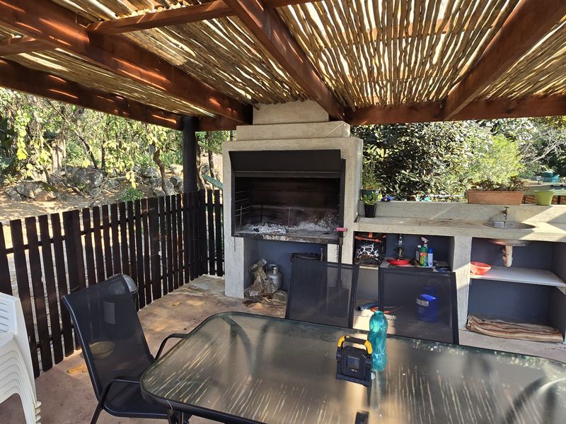 Covered outdoor patio area featuring a built-in braai fireplace, integrated concrete prep counter with sink, and a rustic timber and thatch roof structure.