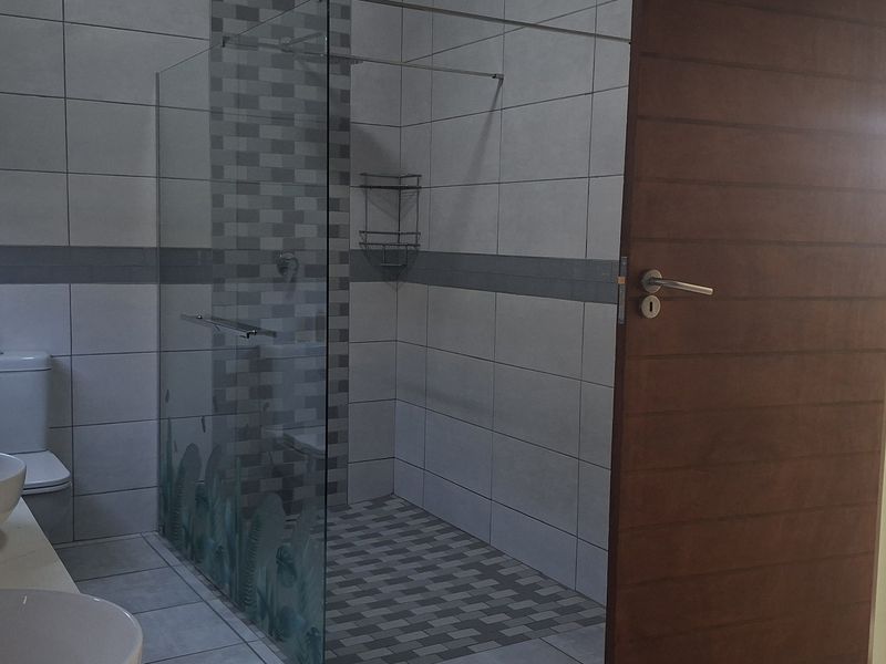 Modern bathroom featuring a double vanity, glass-enclosed shower, and neutral wall tiling, adjacent to an open-plan living area.