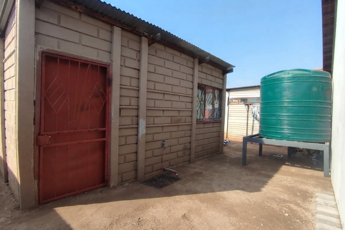 Rear exterior view showing a precast concrete structure with a security gate, corrugated roof, and a large water storage tank on a metal stand.