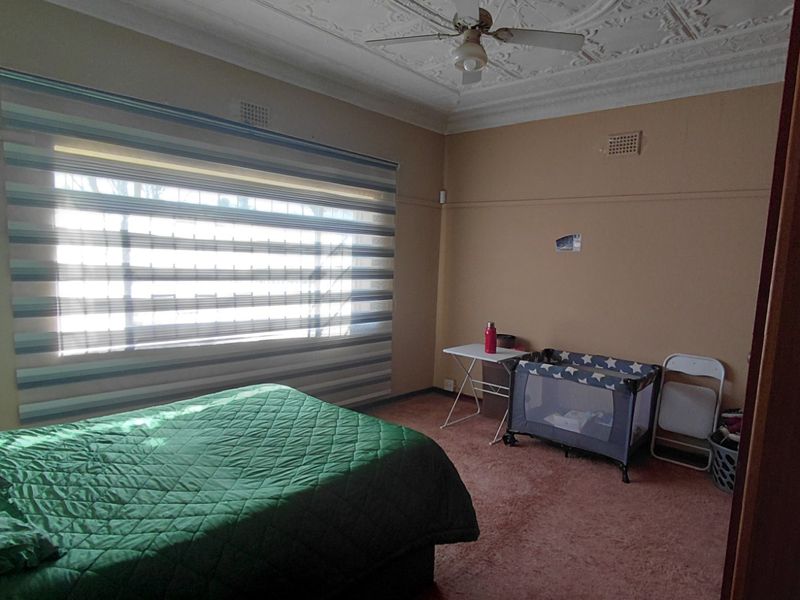 A spacious bedroom featuring a decorative ceiling rose, ceiling fan, and large window with fitted blinds, finished with carpeted flooring.