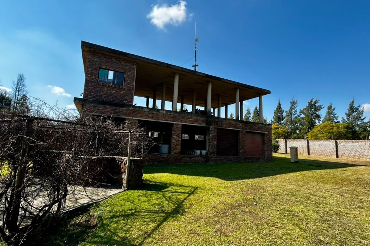 Rear view of a multi-level brick small holding property featuring an expansive upper balcony, integrated garage space, and a large surrounding garden area.