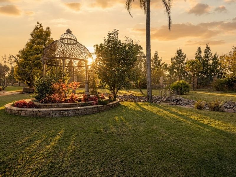 A beautifully landscaped garden featuring a decorative metal gazebo, stone retaining walls, and mature trees, captured during a scenic sunset.