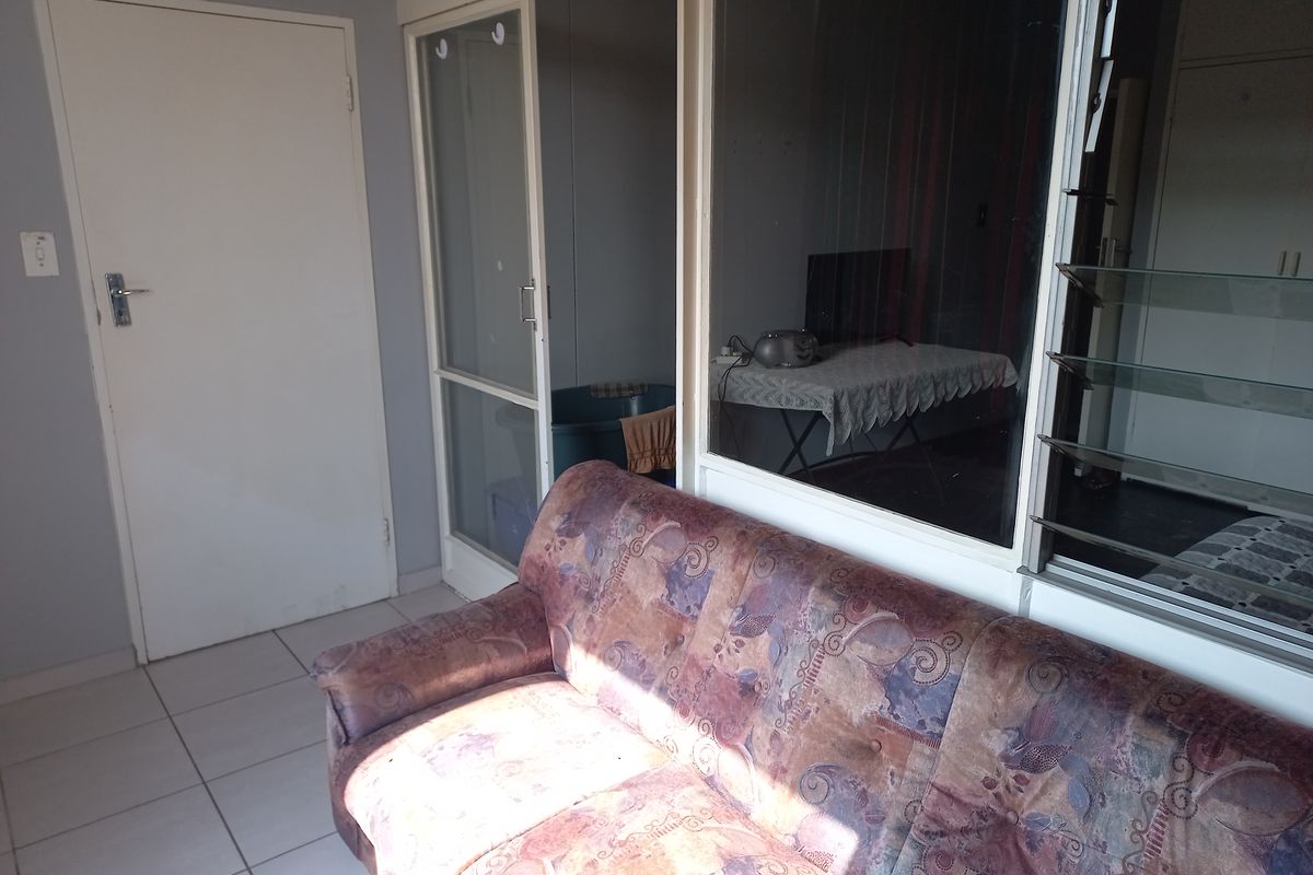 A living area featuring tiled flooring, a sliding glass door, and louvre windows, showing signs of wear and dated finishes.