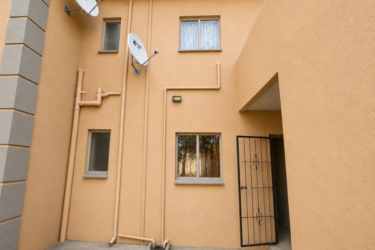Rear exterior view of the apartment featuring a concrete patio, security gate, and visible external plumbing infrastructure.