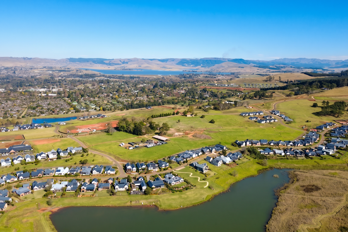 An expansive aerial view of a residential estate featuring a large dam, rolling green hills, and established housing developments under a clear blue sky.