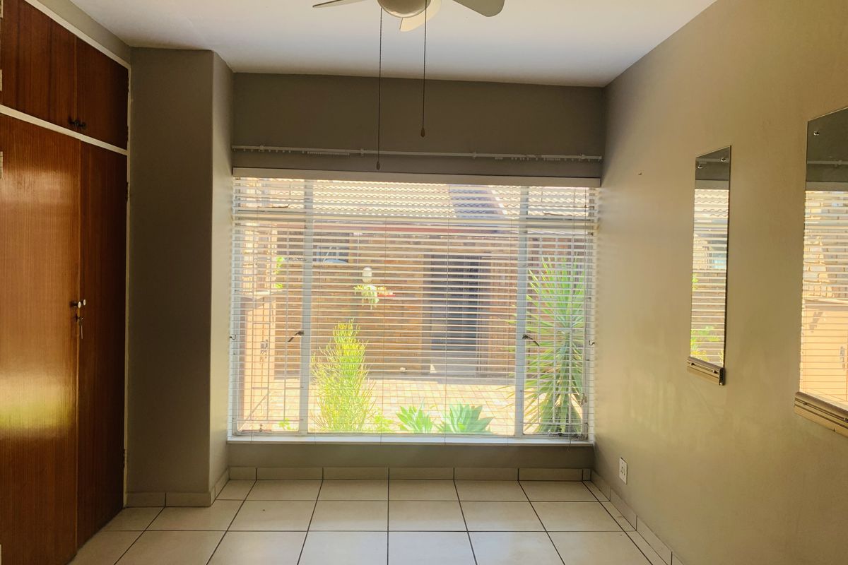 A bright, empty bedroom featuring tiled flooring, built-in wooden cupboards, a ceiling fan, and a large window with venetian blinds overlooking the exterior.