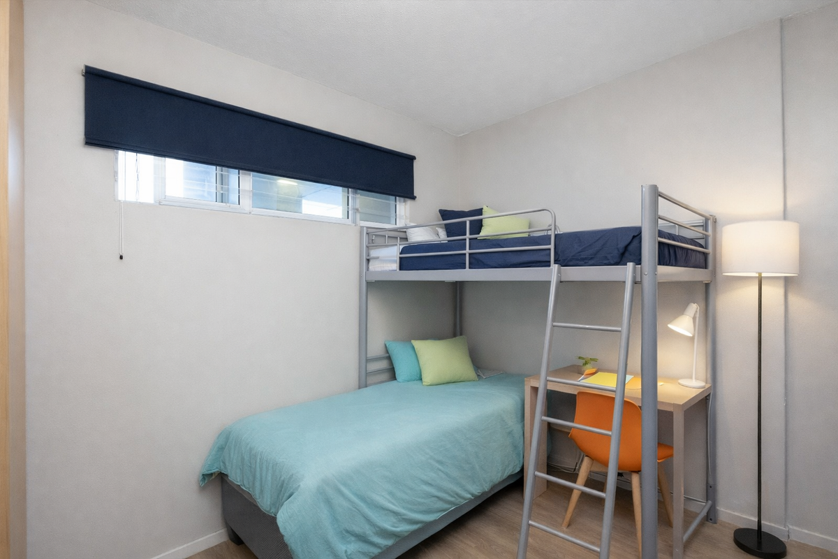 A compact bedroom featuring neutral walls, laminate flooring, and a functional window with a dark roller blind.