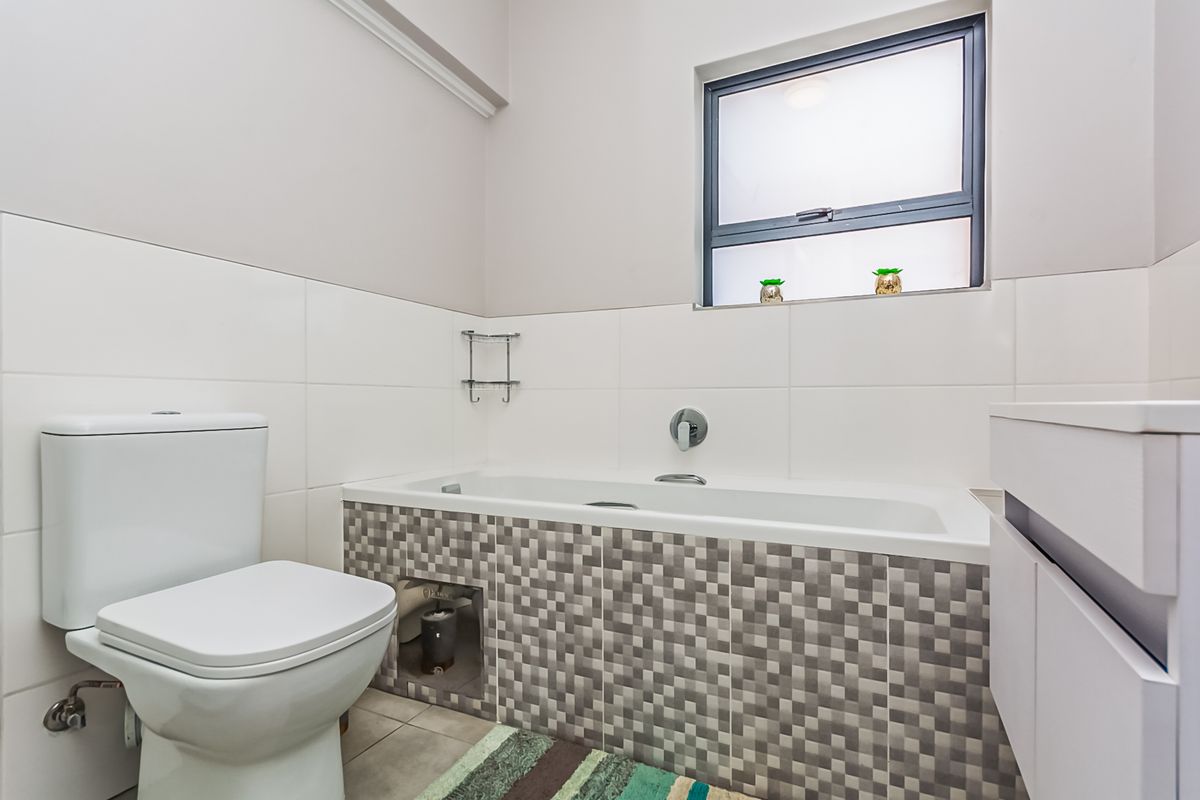A clean bathroom featuring a white bathtub with a mosaic tile apron, a modern toilet, and a vanity unit with a frosted glass window.
