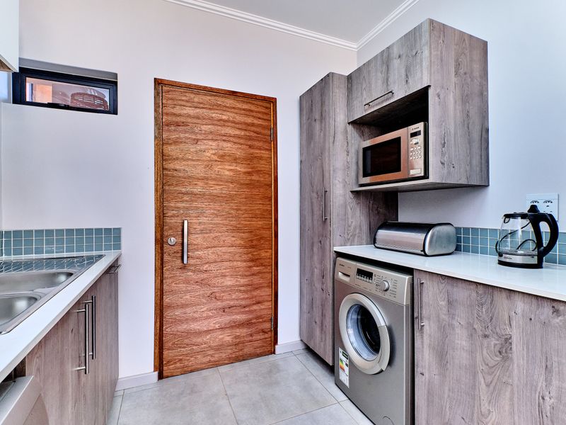 A modern, well-appointed laundry room featuring wood-grain cabinetry, a stainless steel sink, tiled backsplash, and integrated microwave space.